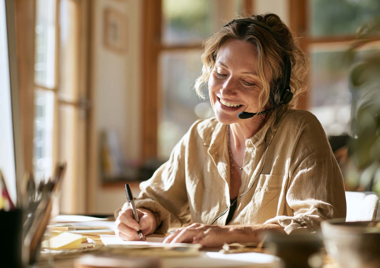Woman wearing a headset smiling while writing notes in a sunlit home office
